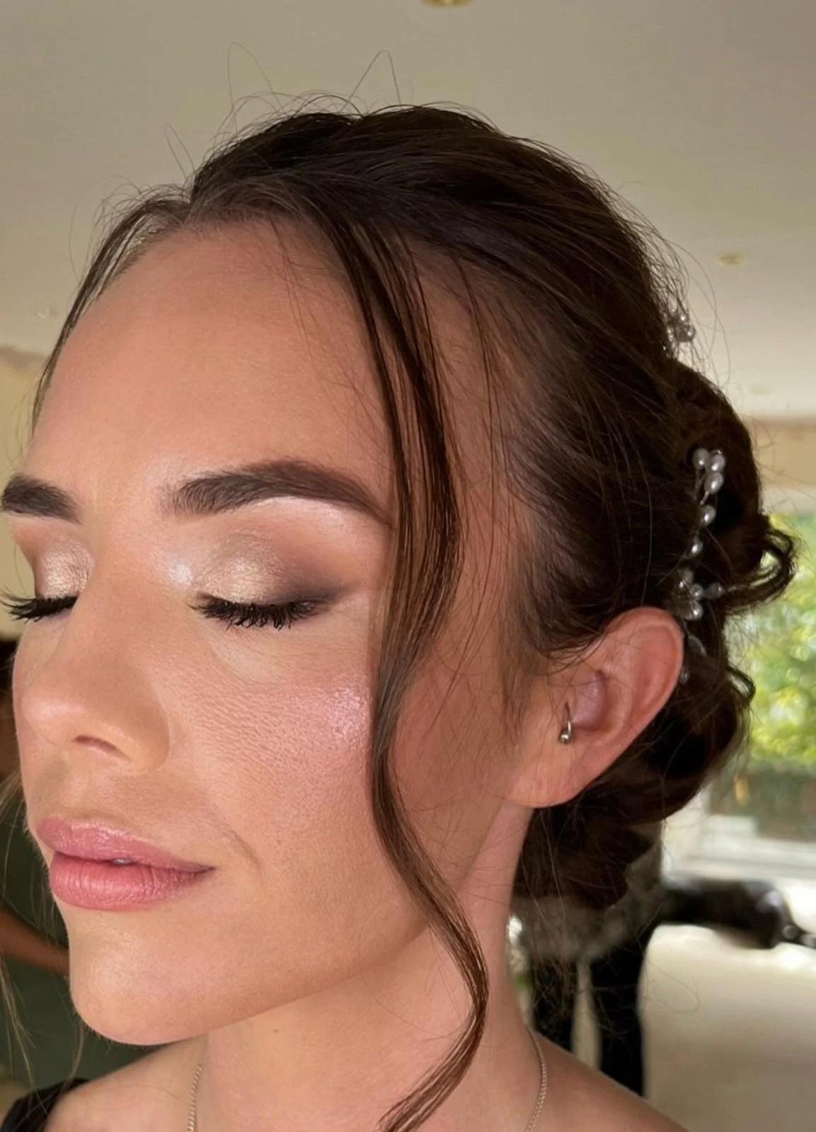 Close-up of a woman's face with makeup, her eyes closed, showing eye shadow, eyelashes, and natural pink lips. Her dark hair is styled up with pearl hair accessories and a loose curl framing her face.