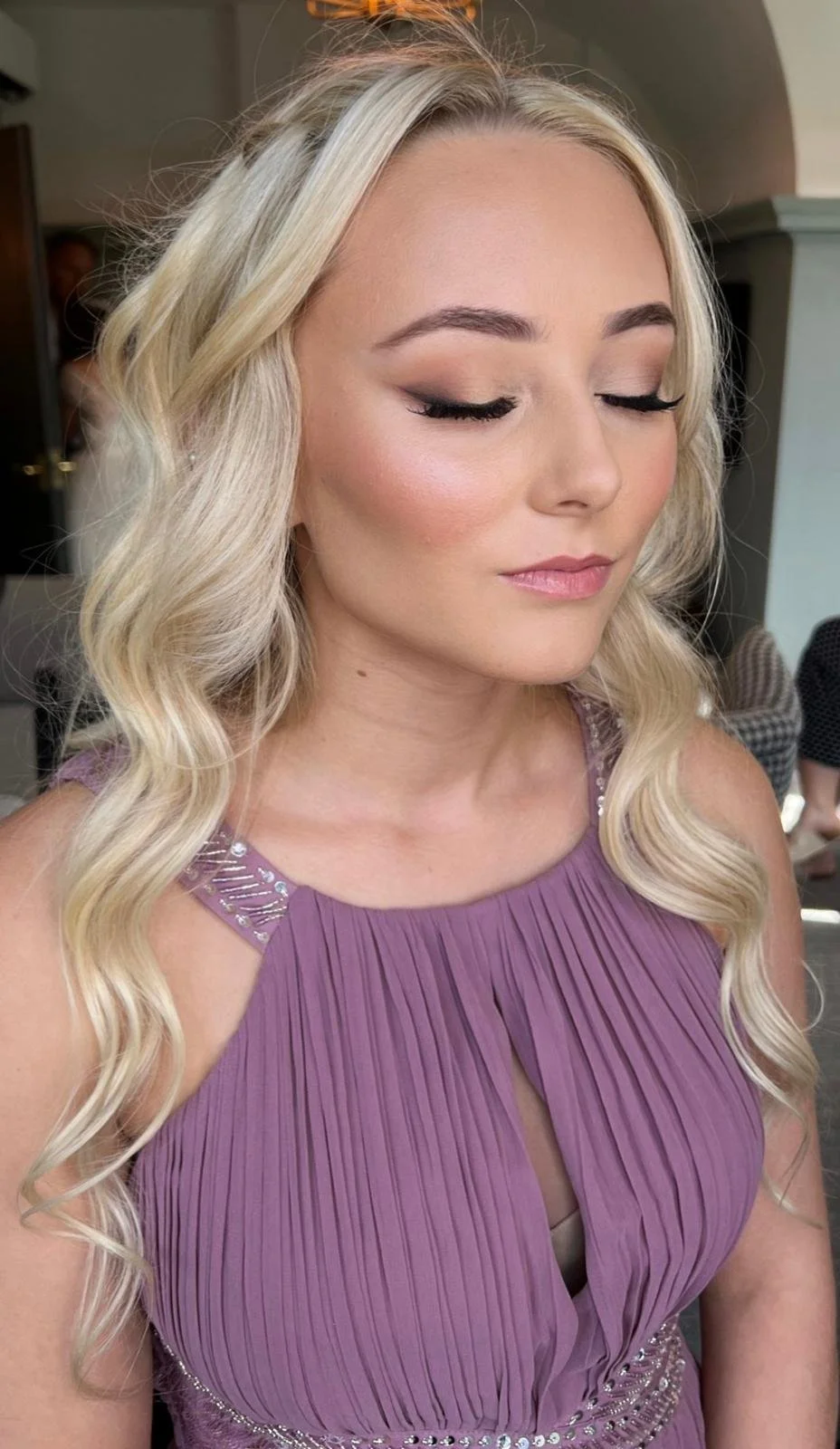 A woman with long, wavy blonde hair wearing a purple pleated dress with embellished shoulder details.