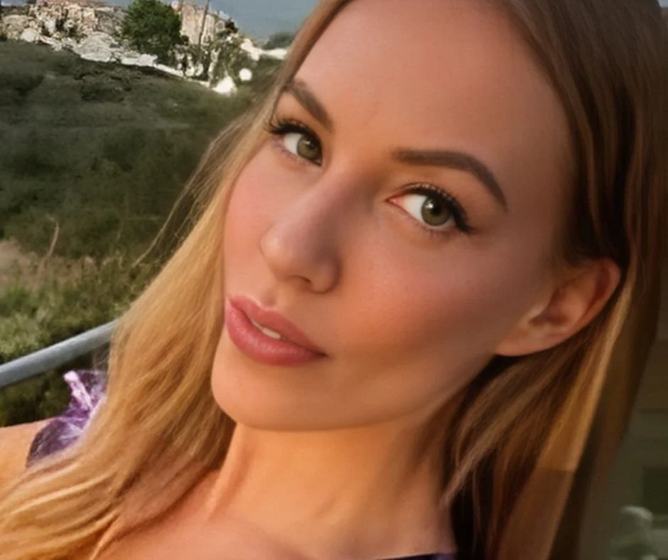 Close-up of a woman with long light brown hair, green eyes, and a slight smile, outdoors near greenery and rocks.