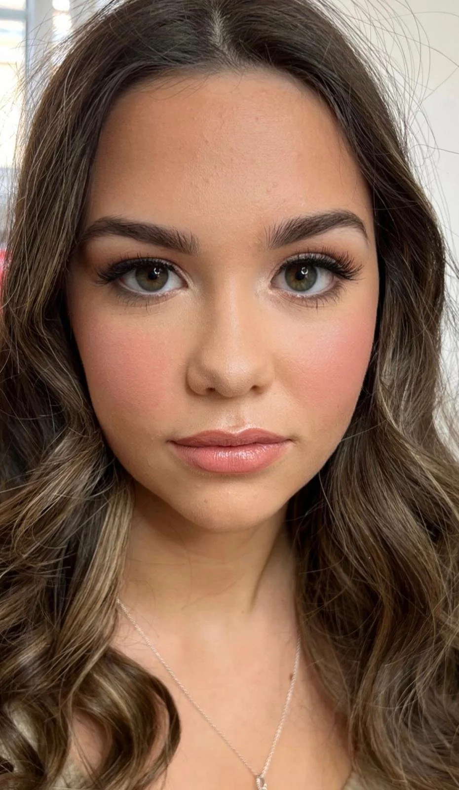 A young woman with long, wavy brown hair, hazel eyes, and makeup including eyeshadow, mascara, blush, and lipstick, wearing a delicate necklace.