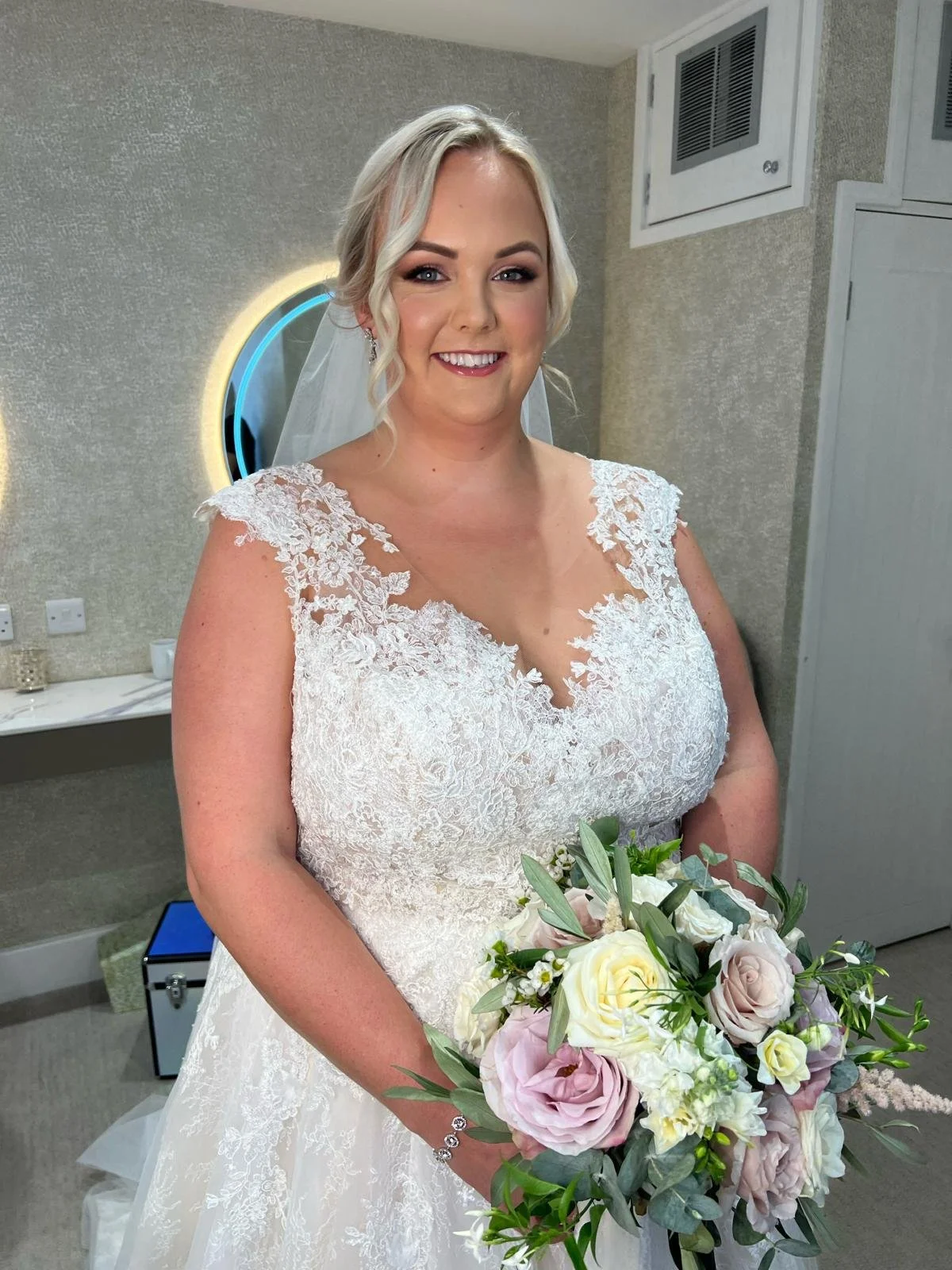 A bride in a lace wedding dress holding a bouquet of roses and greenery, smiling in a room with a mirror and neutral decor.