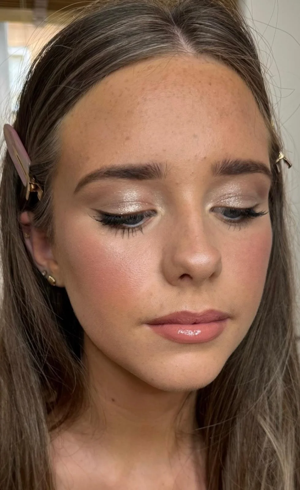 Close-up of a woman with medium-length brown hair, styled with hairpins, wearing makeup that includes shimmery eyeshadow, winged eyeliner, mascara, blush, and nude lipstick.
