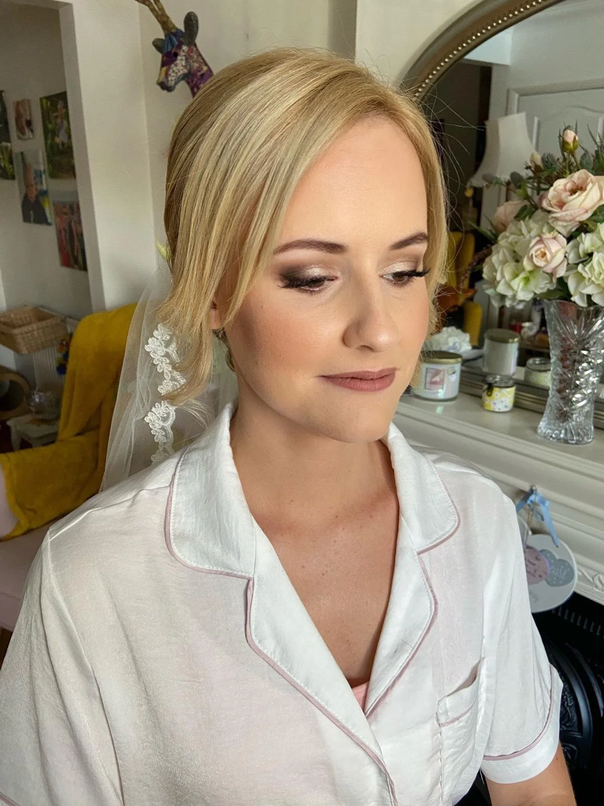 A woman with blonde hair and makeup, wearing white pajamas with pink piping, is indoors in front of a mirror and flowers. She is preparing for a special occasion, possibly a wedding.