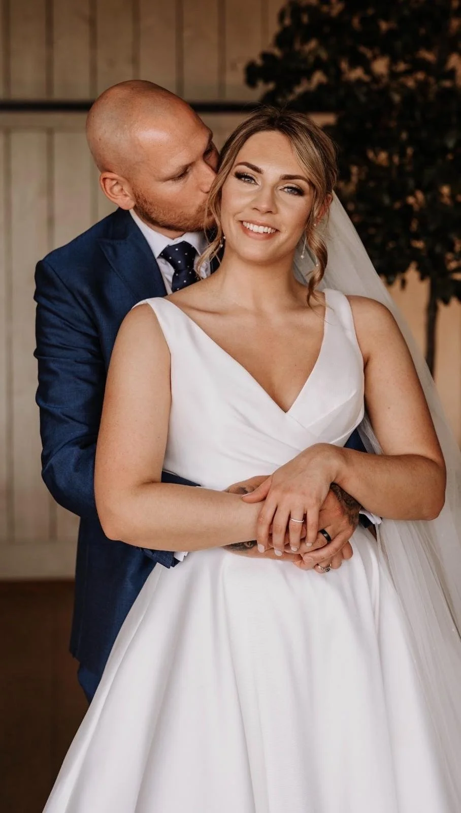 A bride and groom on their wedding day, the groom with a shaved head and wearing a blue suit, is kissing the bride on her cheek. The bride has light brown hair styled in loose waves, and is wearing a white wedding dress with a V-neck. They are smiling and embracing, with a rustic background and a tree in the distance.