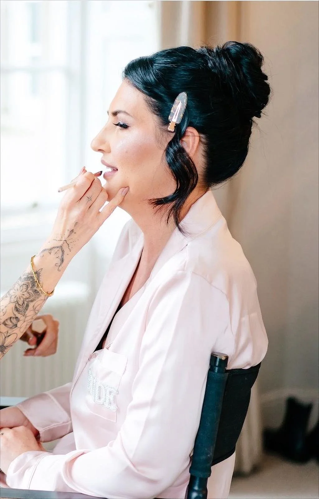 A woman with dark, curled hair secured with a hairclip, sitting on a black chair, getting her makeup done by a makeup artist with tattoos on her arm, in a well-lit room.
