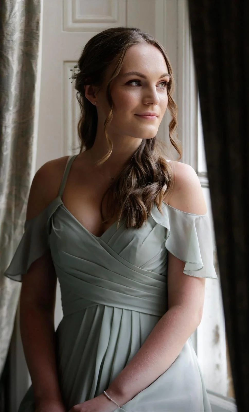 A beautiful bridesmaid with long, wavy, brown hair looking out a window, wearing a sage green off-the-shoulder dress - Makeup by Kelly Marie