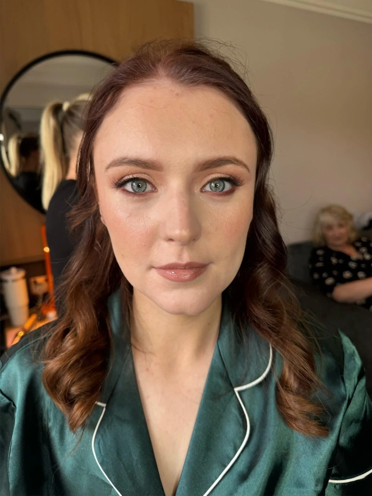 A woman with brown hair and blue eyes wearing green satin pajamas with white piping, looking directly at the camera in a room. In the background, there is a wall mirror, a woman sitting on a couch, and casual room decor.