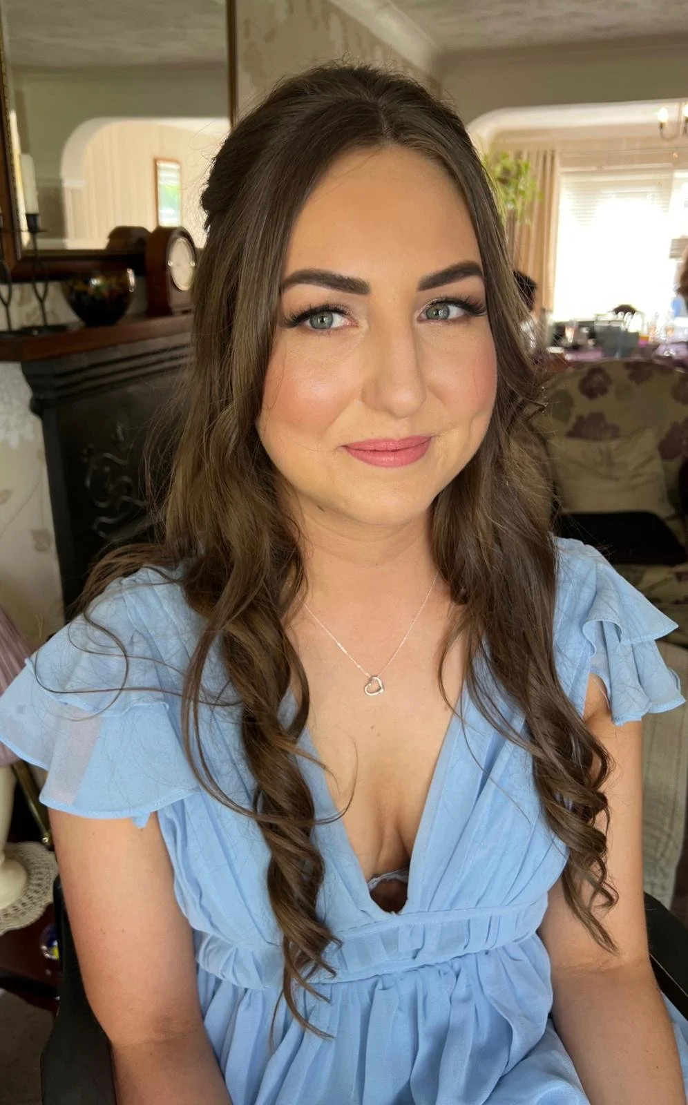 A woman with long brown hair and blue eyes smiling, wearing a light blue dress and a silver heart necklace, sitting indoors in a well-lit room.