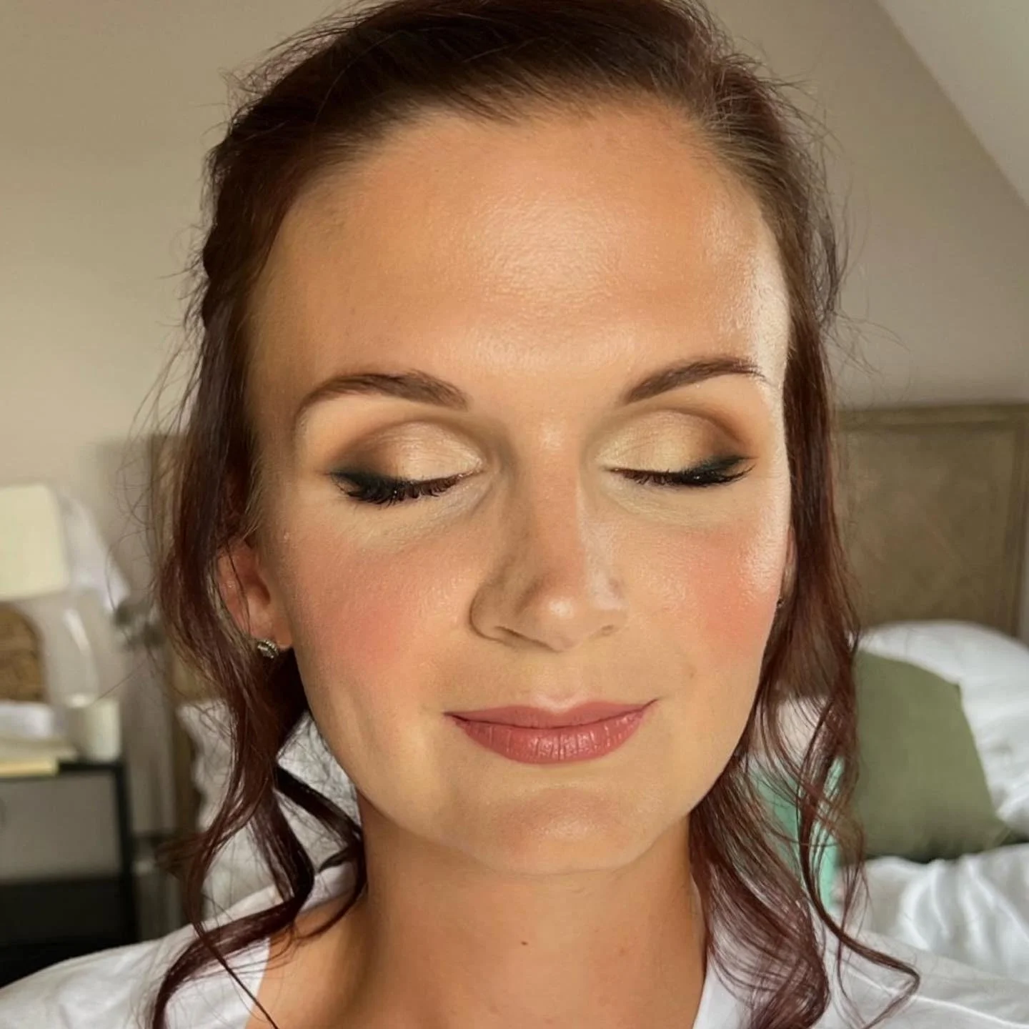 Close-up of a woman's face with makeup, eyes closed, smiling slightly, with brown hair and earrings, in a bedroom.