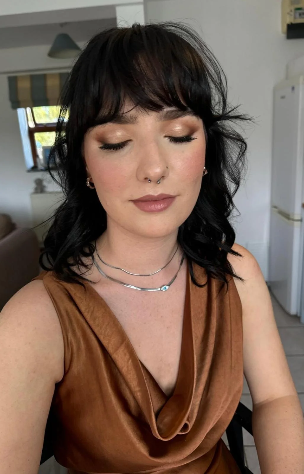 A woman with black hair, closed eyes, wearing makeup, a septum piercing, necklaces, and a brown sleeveless top, sitting in a room with a window, refrigerator, and a gray lamp in the background.