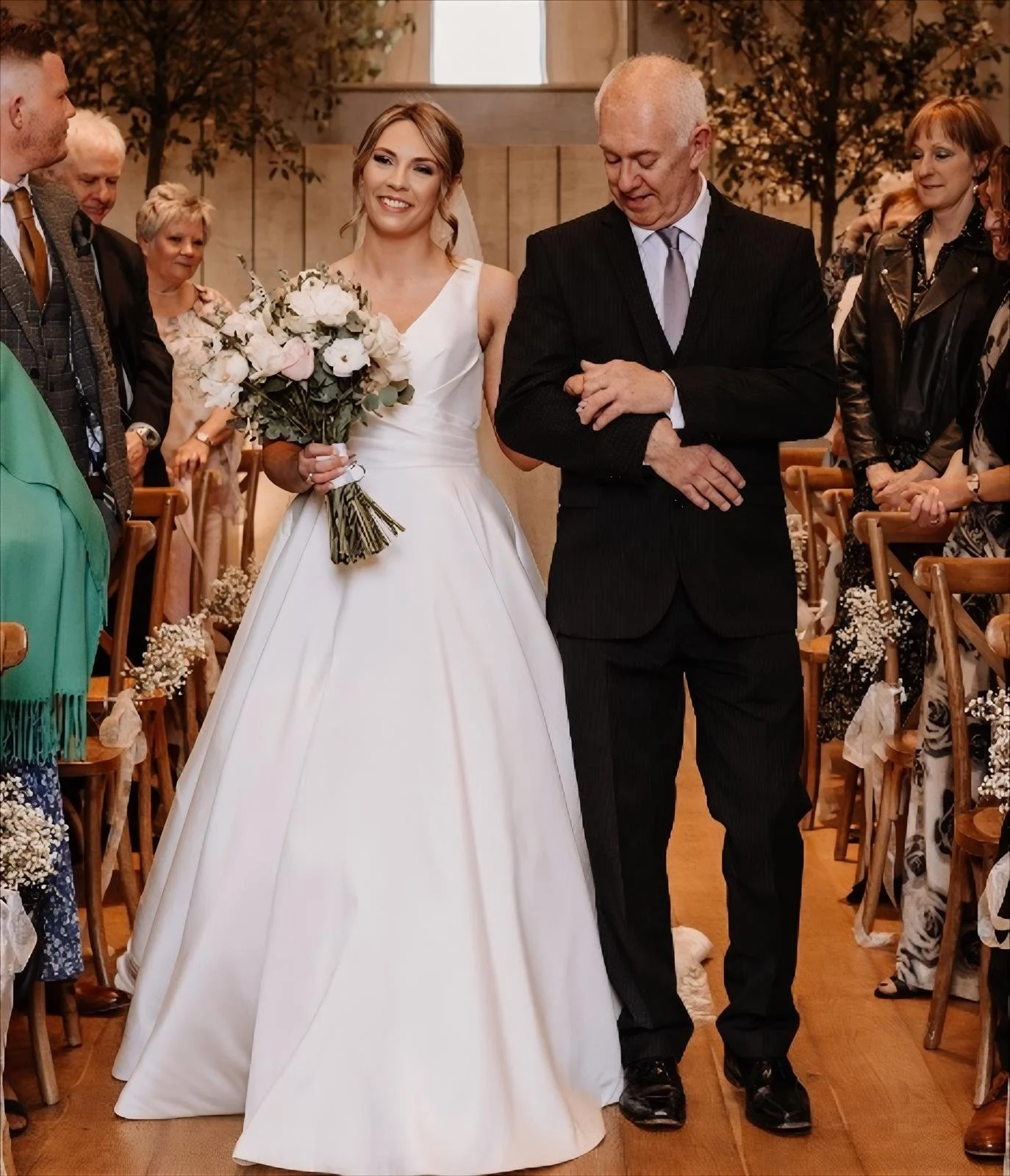 A bride in a white wedding dress holding a bouquet, walking down the aisle with her father, surrounded by wedding guests.