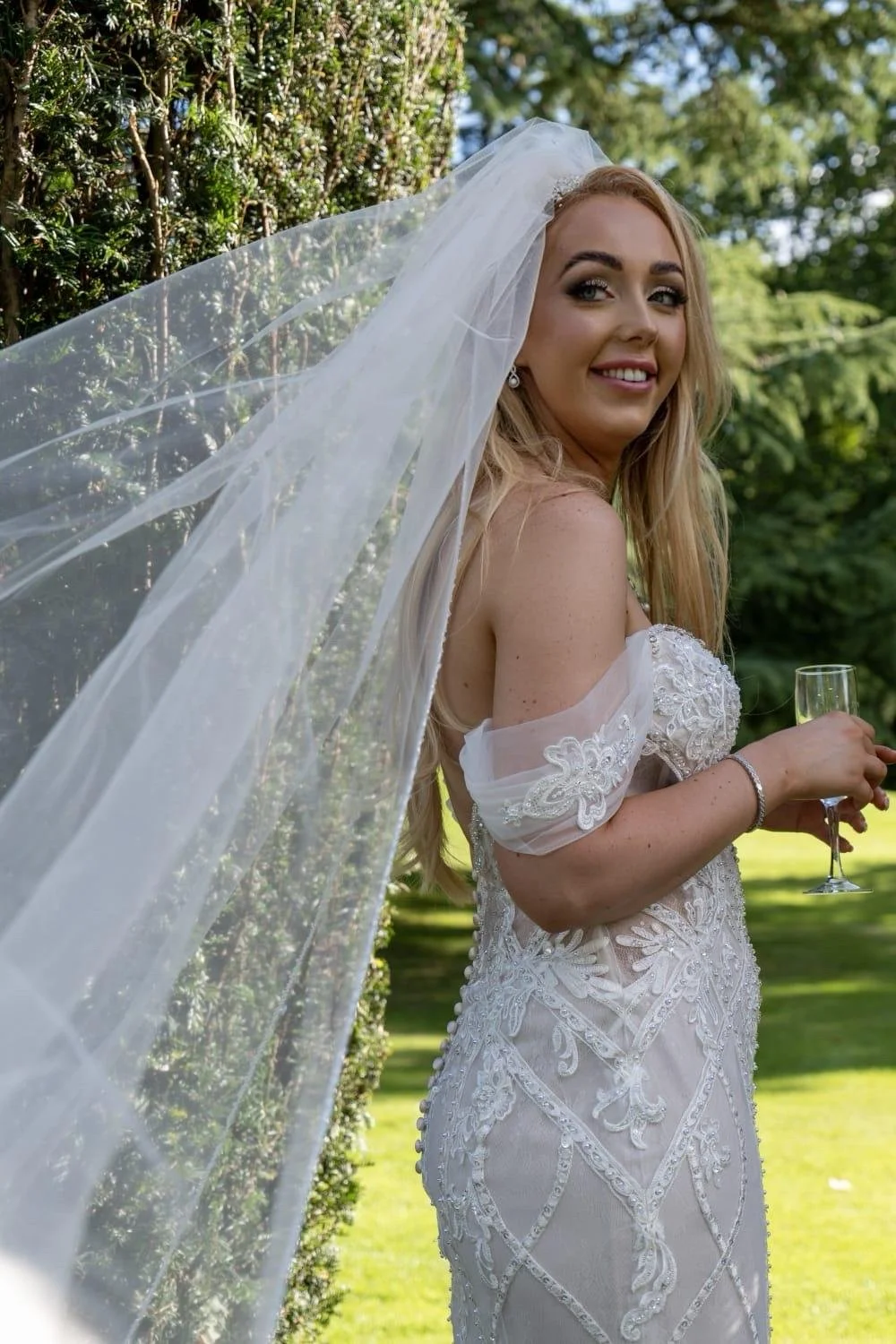 A woman in a wedding dress holding a glass of champagne outdoors with green trees and grass in the background.