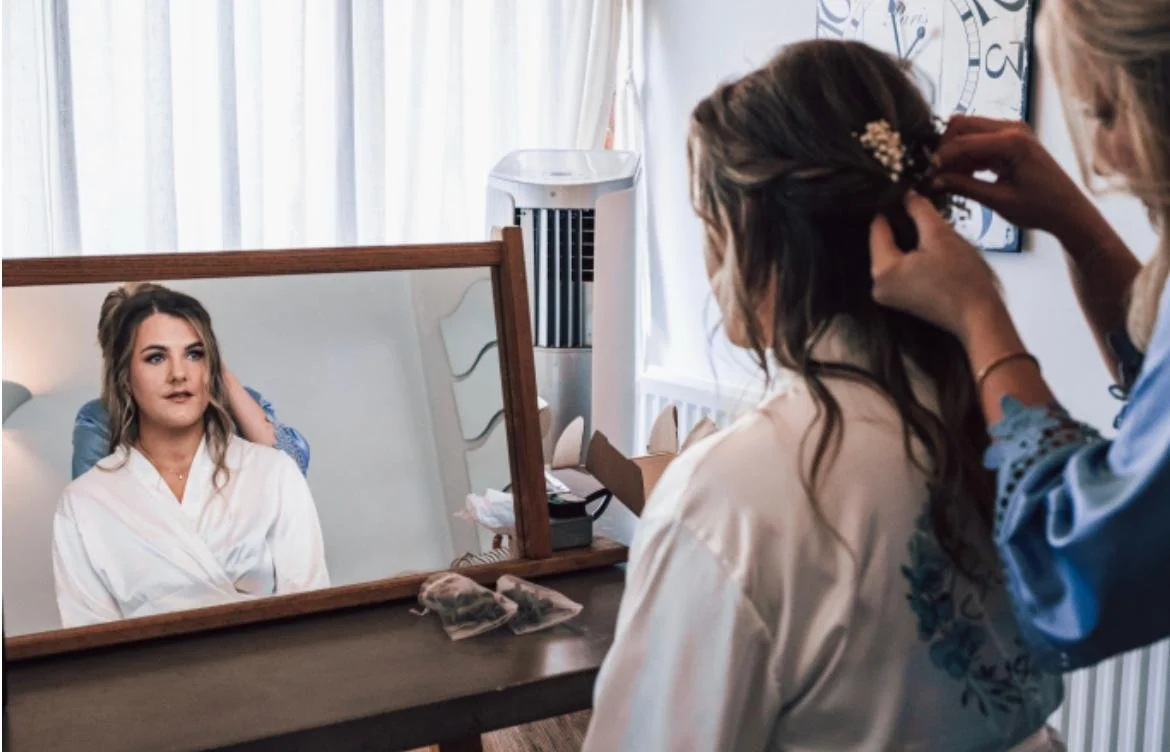 A woman in a white robe is sitting in front of a mirror, getting her hair styled by another woman in a blue top. The mirror reflects her face and the hairstylist working on her hair.