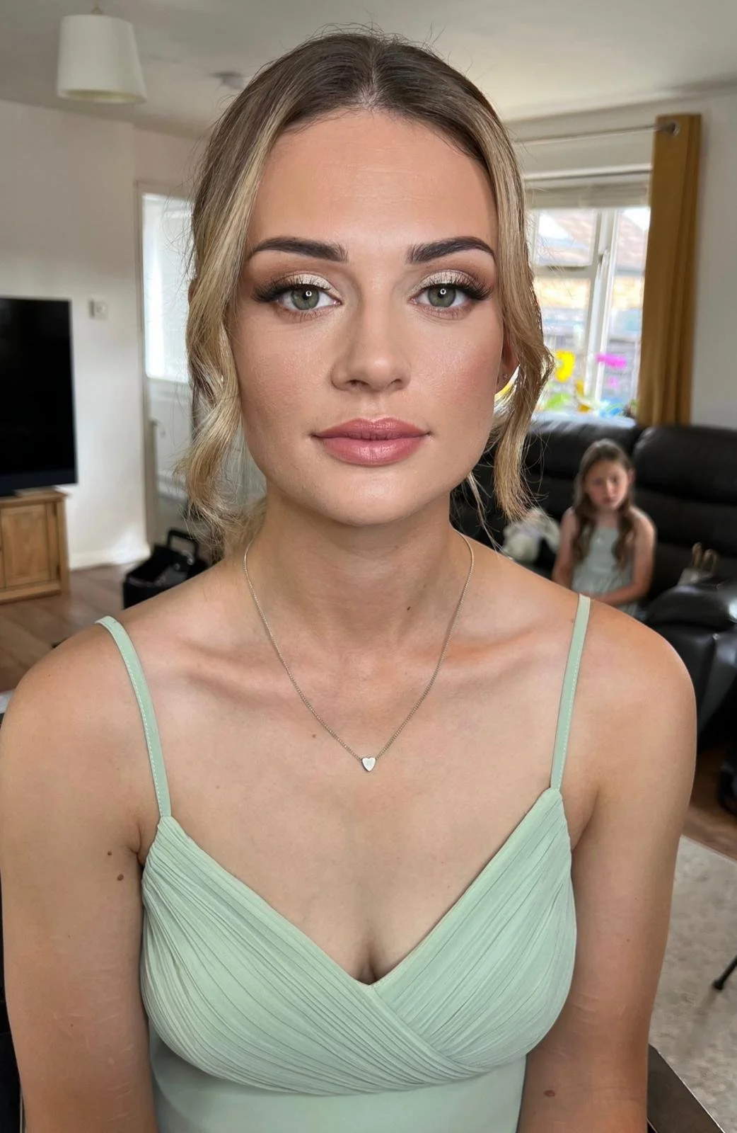 A young woman with blonde hair and blue eyes looking directly at the camera, wearing a light green dress and a silver heart necklace, in a living room with a television and a window in the background, with a young girl sitting on a sofa in the backgr