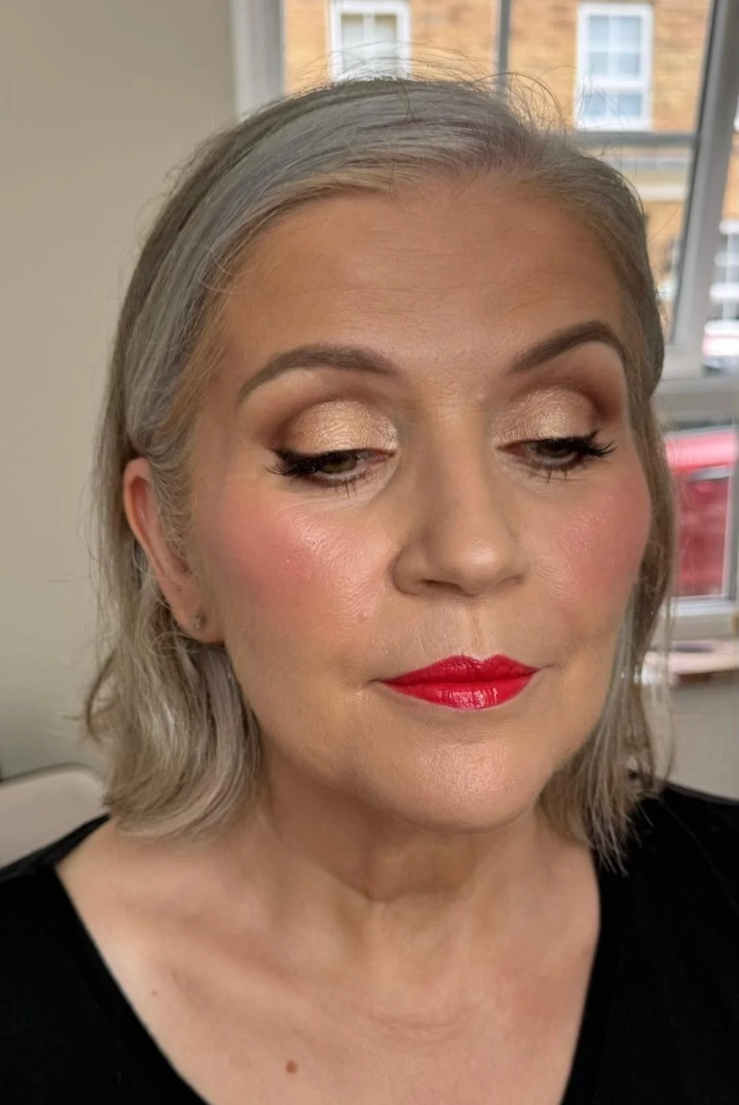 A woman with gray hair, wearing makeup including gold eyeshadow and red lipstick, with her eyes closed.