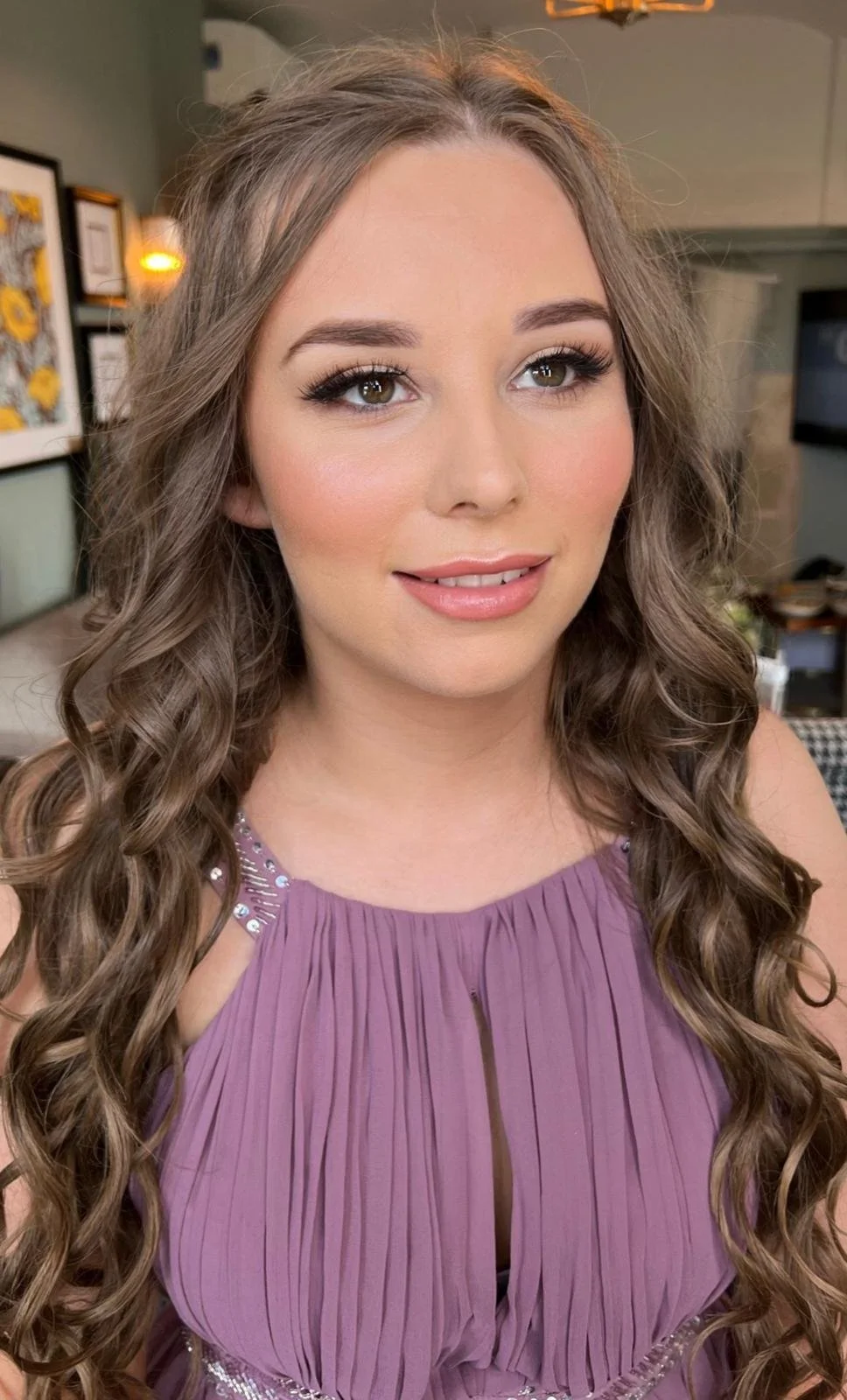 Close-up portrait of a young woman with long, curly brown hair, wearing a purple pleated dress with embellishments on the shoulders, in a well-lit room with artwork and a TV in the background.