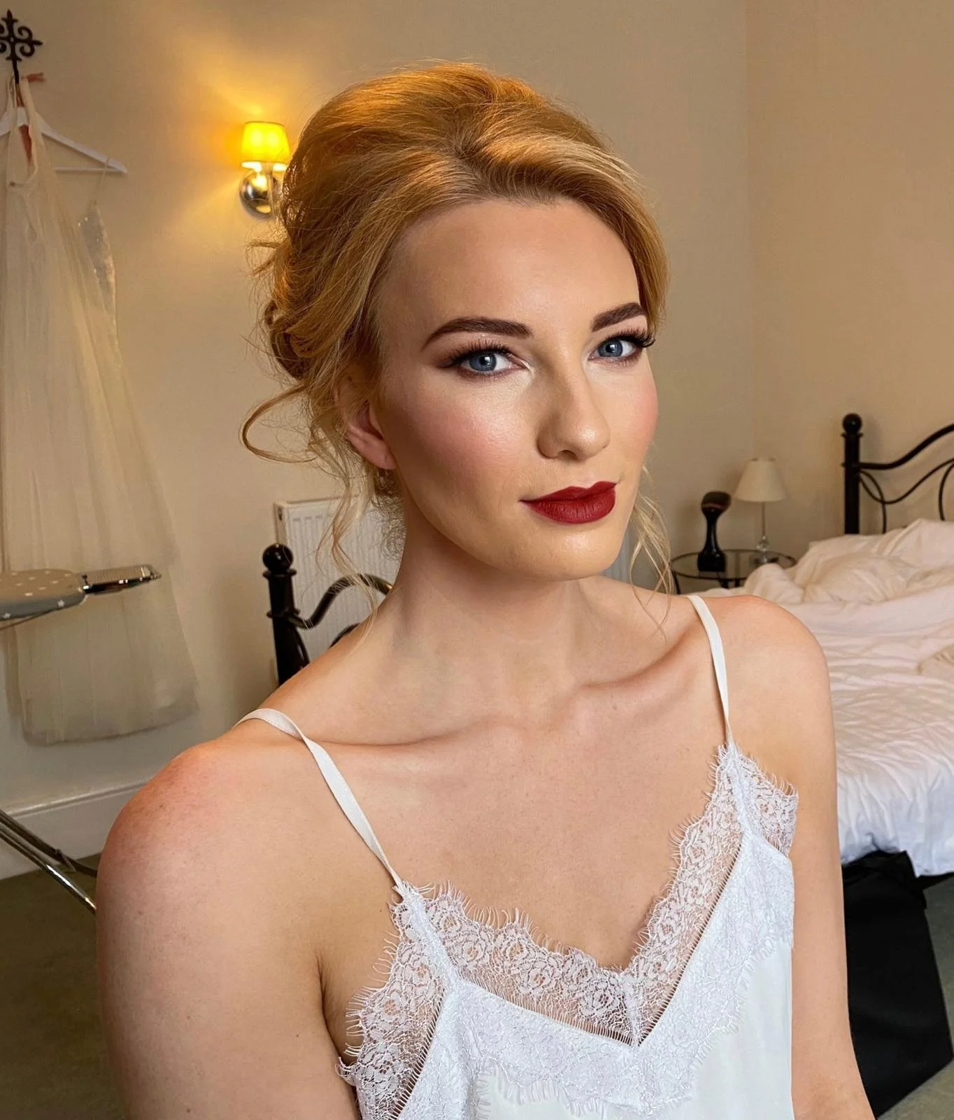 A woman with blonde hair styled in an elegant updo, wearing makeup with red lipstick and blue eyes, dressed in a white lace camisole, in a bedroom with a bed, bedside lamps, and a wall-mounted light