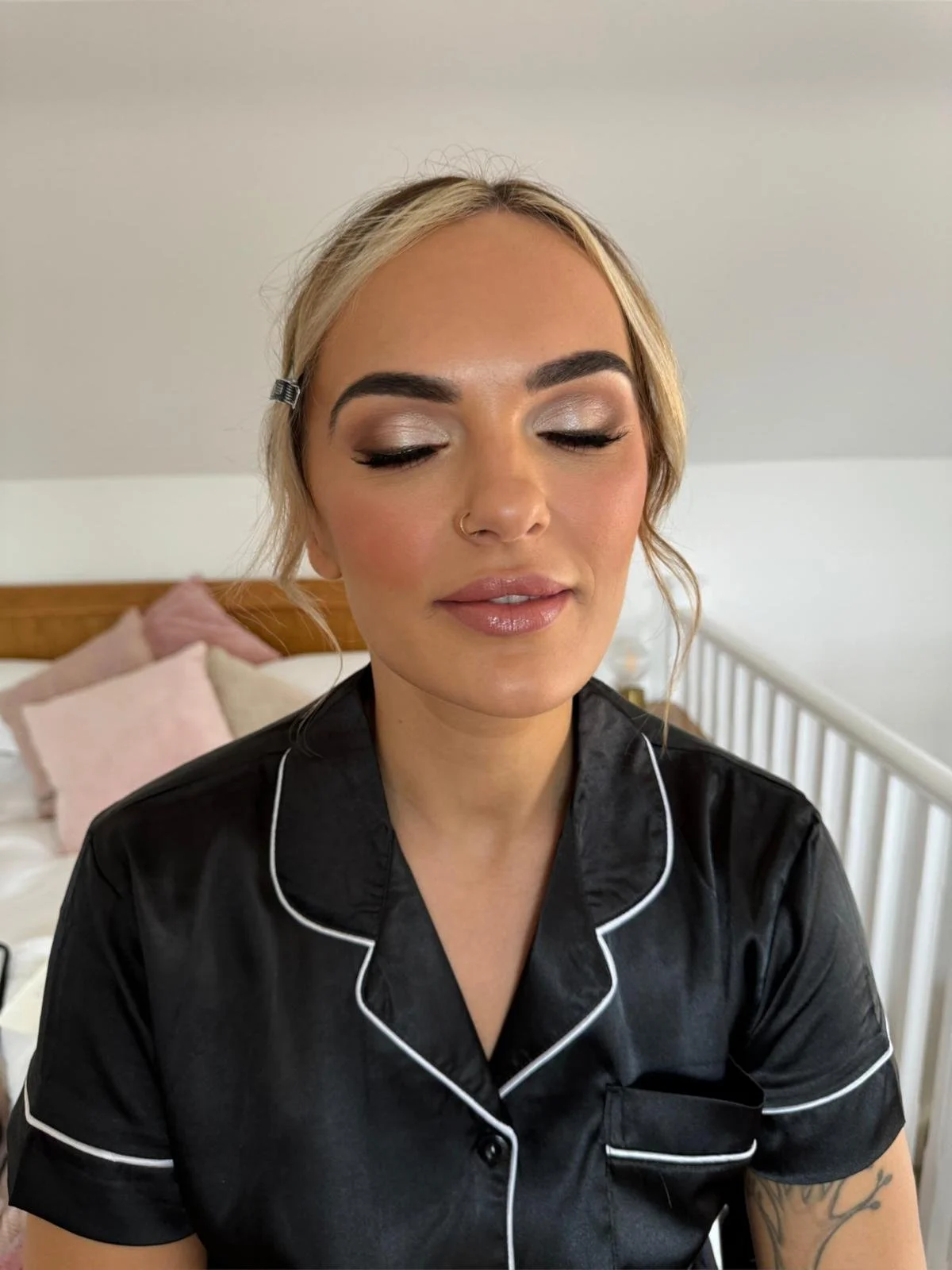 A woman with blonde hair, styled with side part and hair clip, wearing makeup with silver eyeshadow, long eyelashes, and nude lipstick, dressed in black satin pajamas with white piping, sitting on a bed with white railing and pink and white pillows i