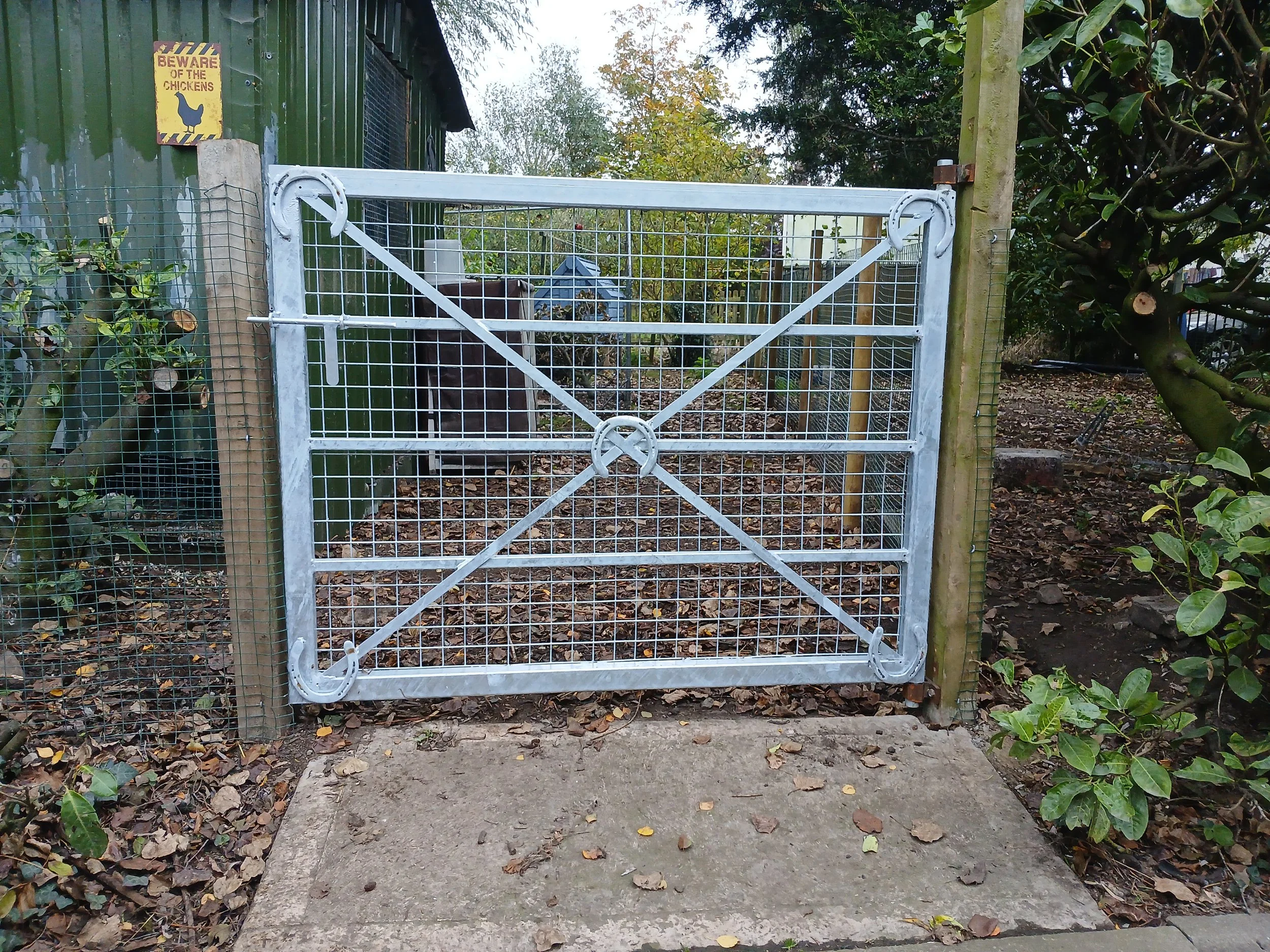 A metal gate with a cross brace in a garden, connected to wooden posts with mesh fencing on the sides, surrounded by green plants and trees.