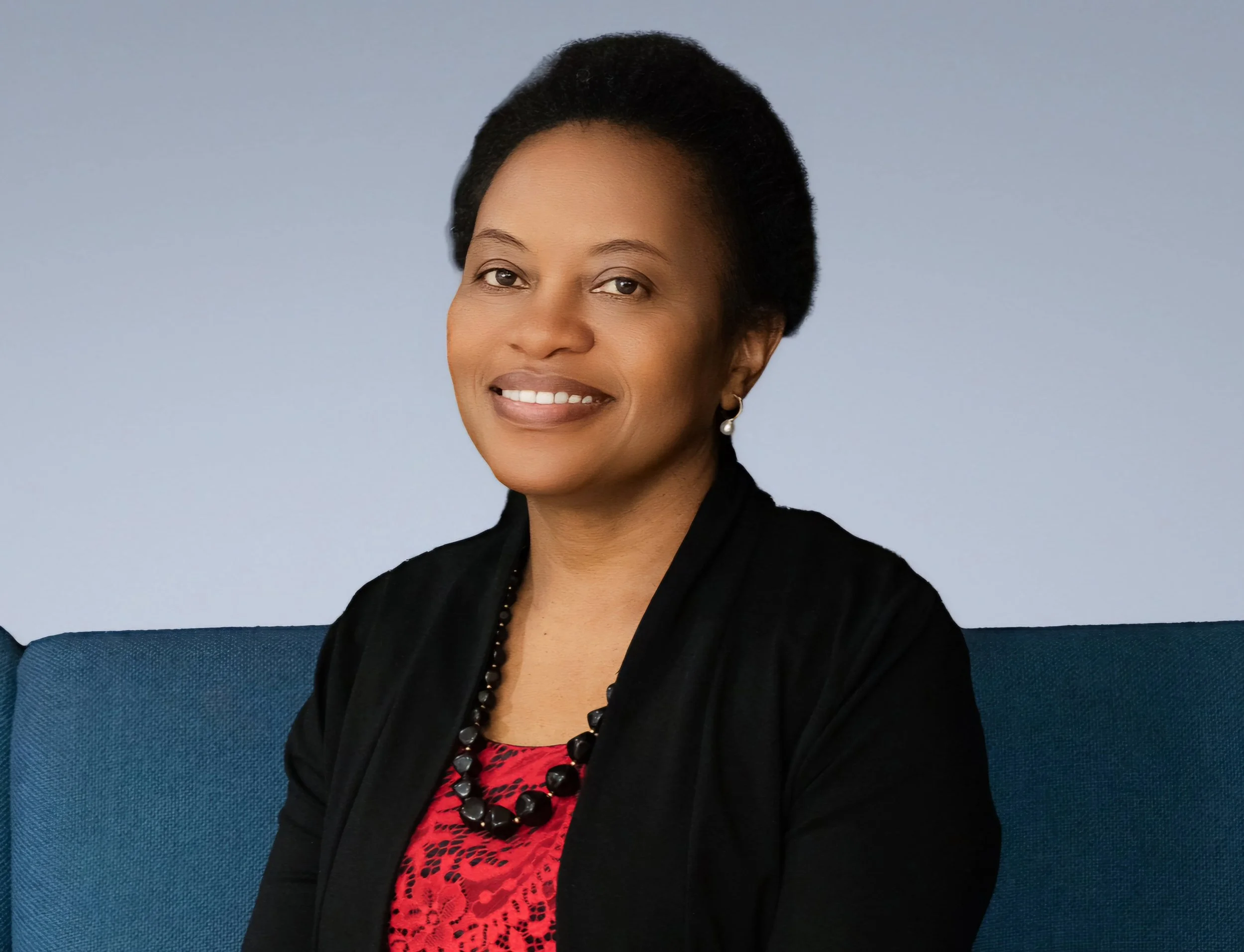 Portrait of a woman with short black hair, wearing a black jacket, red patterned blouse, pearl earrings, and a black beaded necklace, sitting on a blue couch against a light blue background.