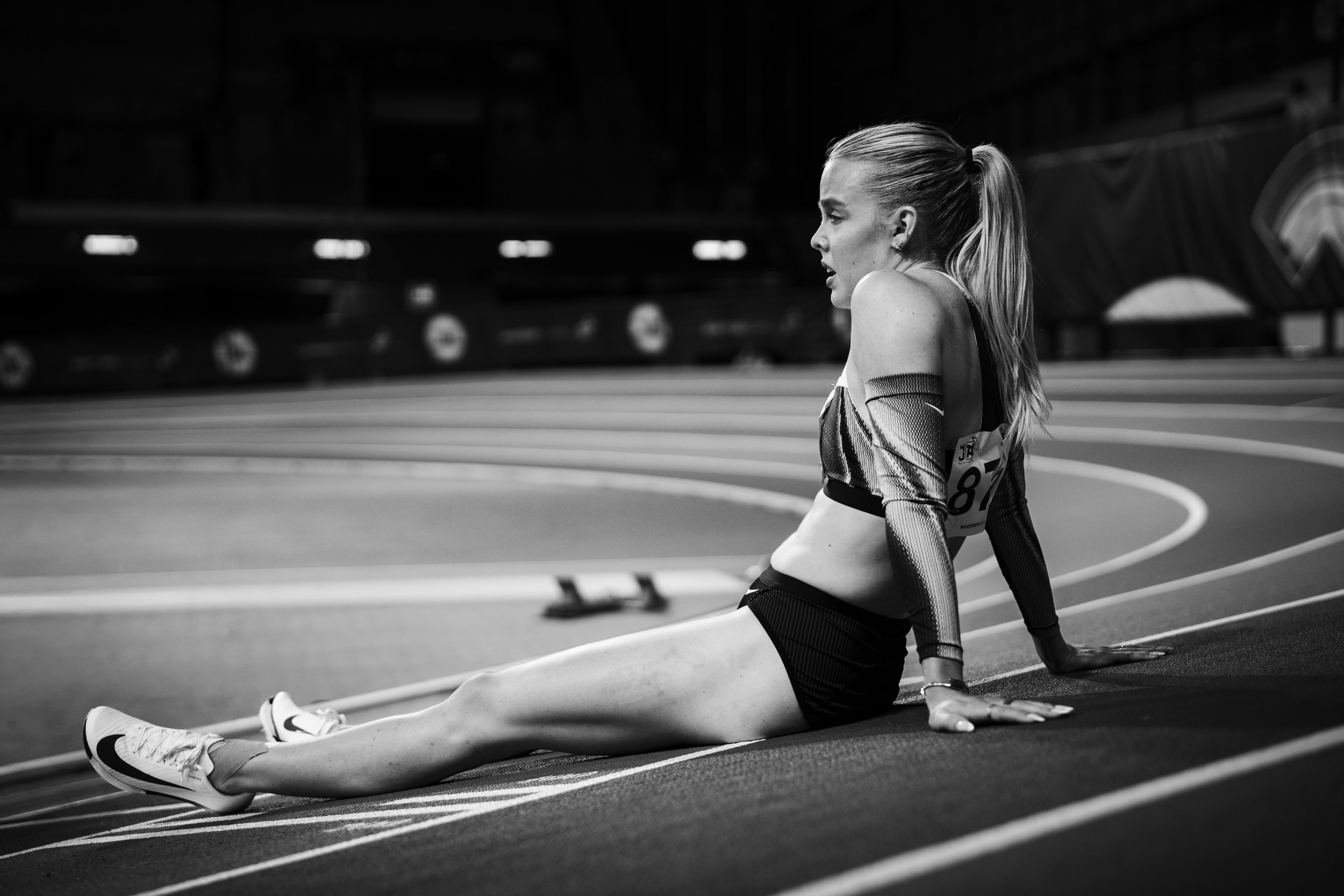 A female athlete sitting on a track in a sports stadium, resting after running, wearing athletic clothing and sneakers.