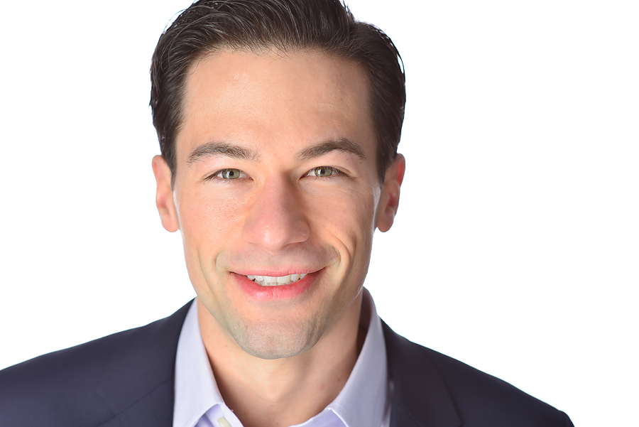 Close-up of a smiling man with dark hair, wearing a suit and white shirt, against a white background.