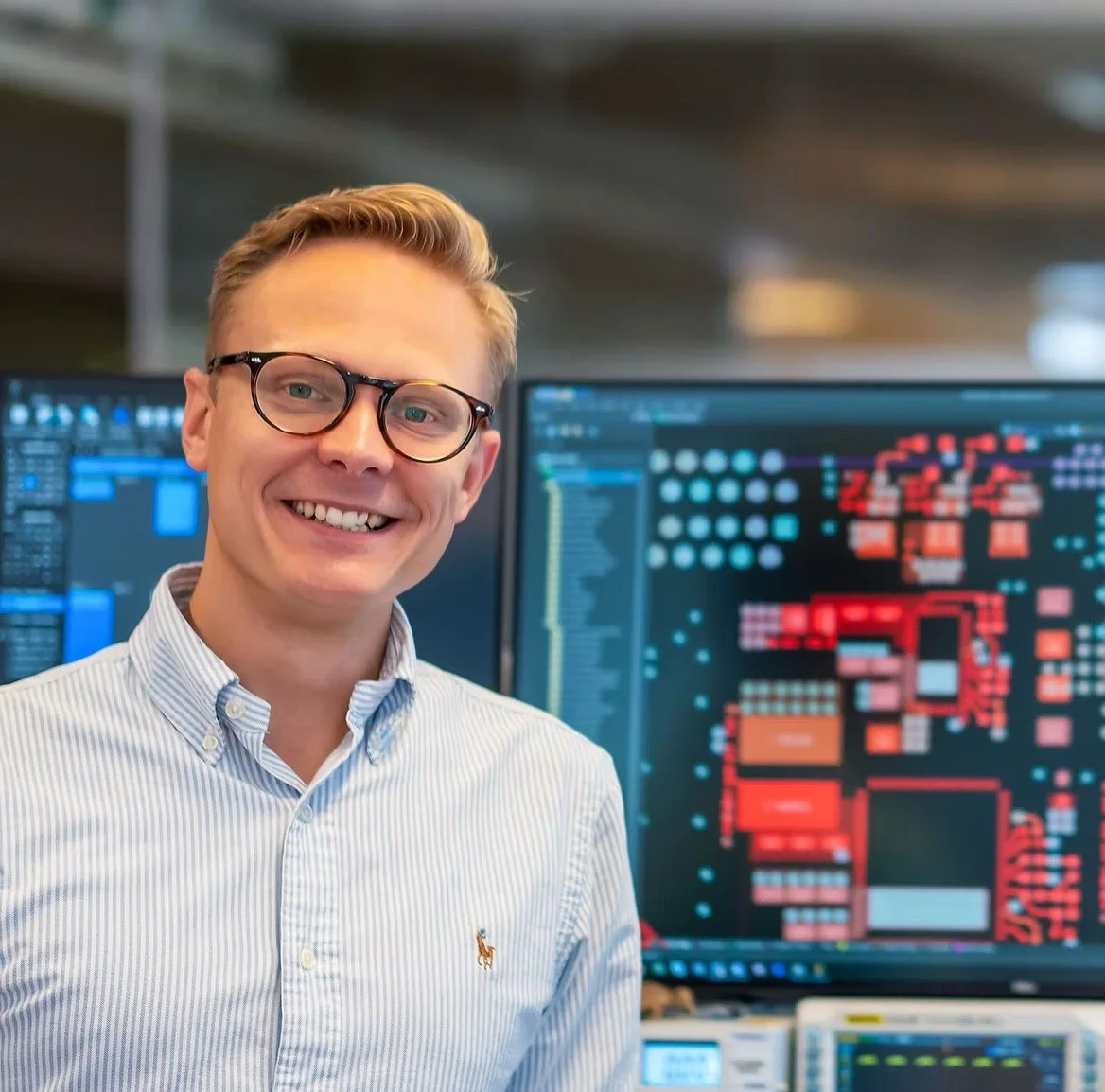 A smiling man wearing glasses and a light blue button-up shirt stands in front of computer screens displaying circuit board diagrams and electronic schematics.