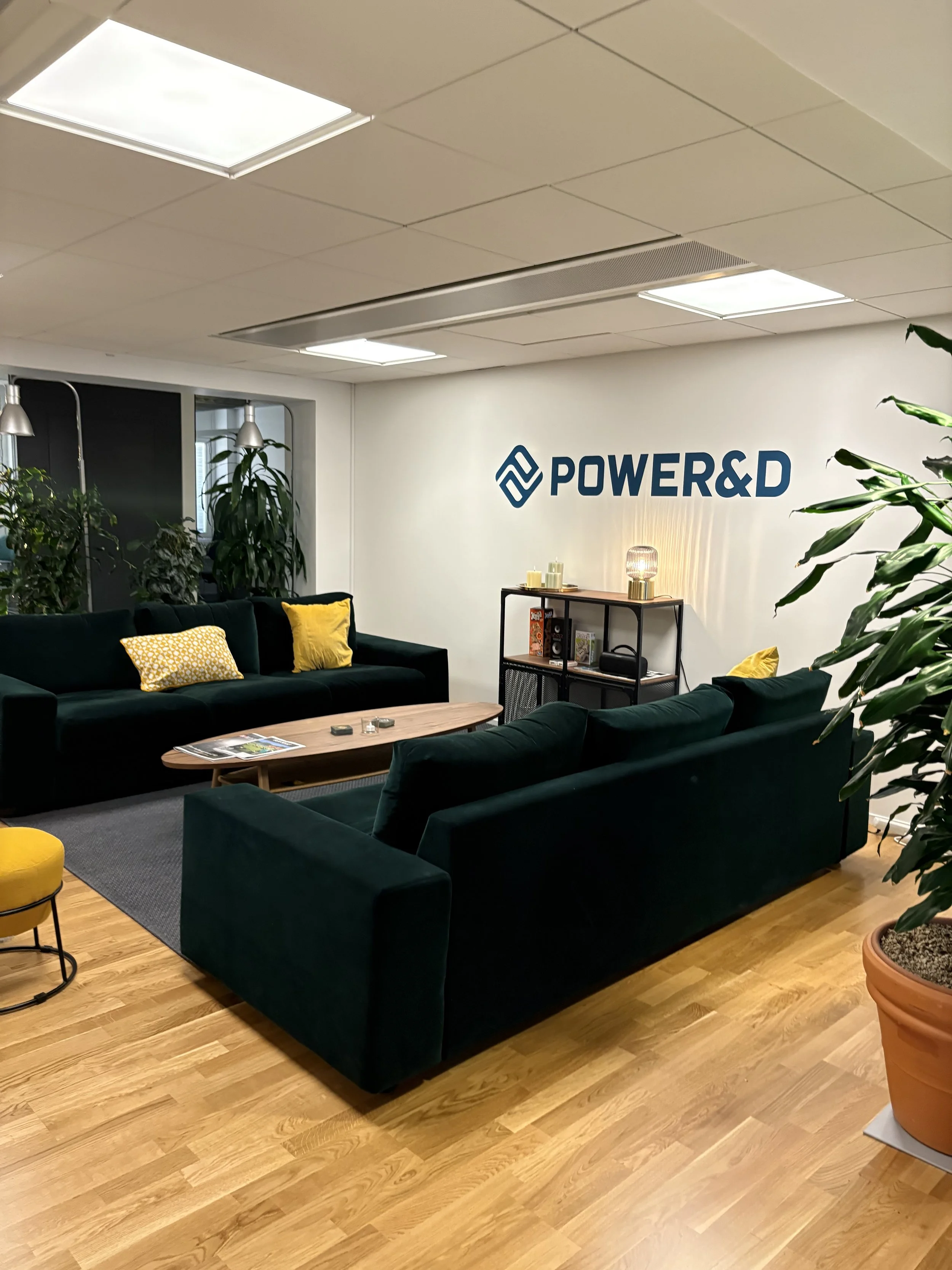 An office lounge area with dark green sofas, yellow pillows, a wooden coffee table, potted plants, and a wall with the logo "POWER&D"