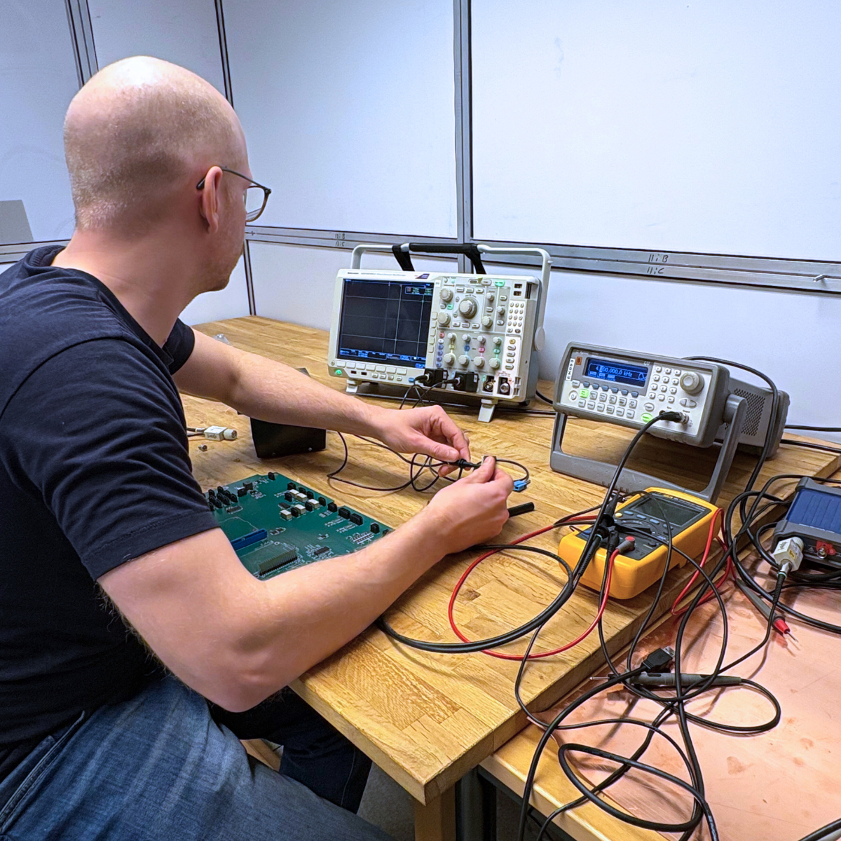 A man with glasses working at a wooden table with electronic testing equipment, including oscilloscopes, a multimeter and a circuit board, inside a laboratory.