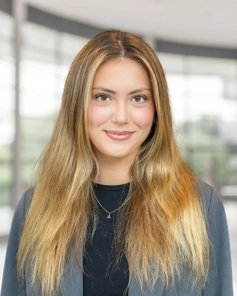 A young woman with long, wavy, light brown hair and fair skin, smiling, wearing a black top and a black blazer in a modern indoor setting with large windows.