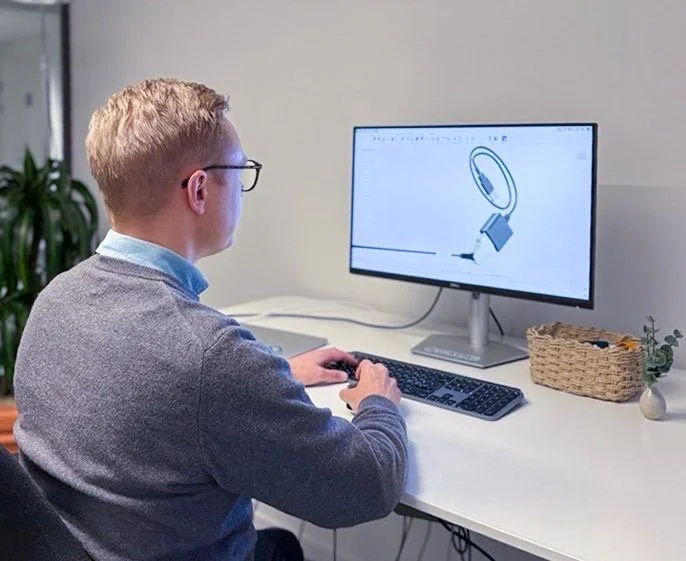 A man using a computer with a 3D design of a calve on the screen. He is sitting at a white desk with a keyboard, mouse and a small basket and plant.