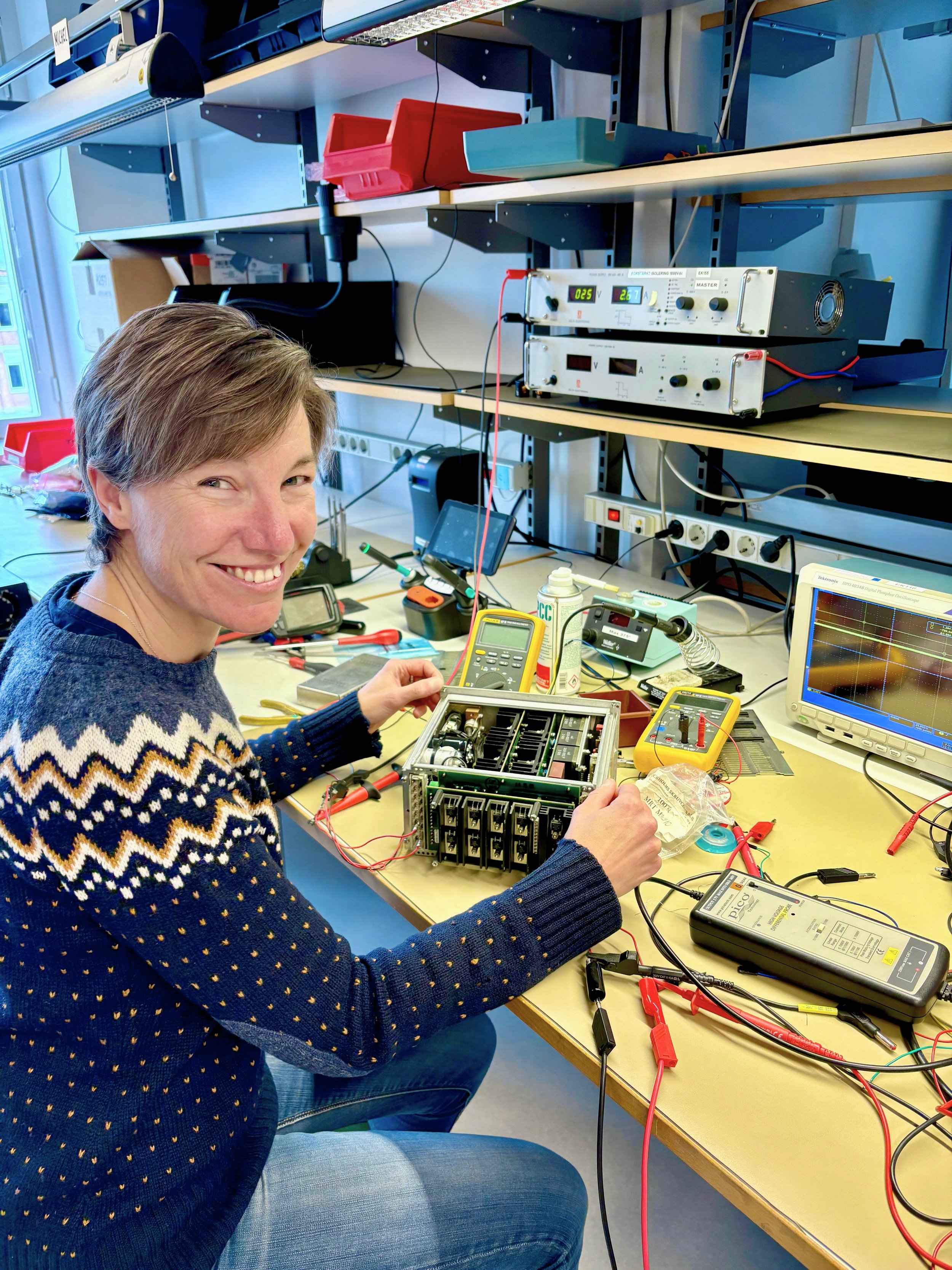A woman in a blue patterned sweater working on electronic equipment at a laboratory bench with test instruments and circuit boards,  surrounded by electronic testing and measurement devices.