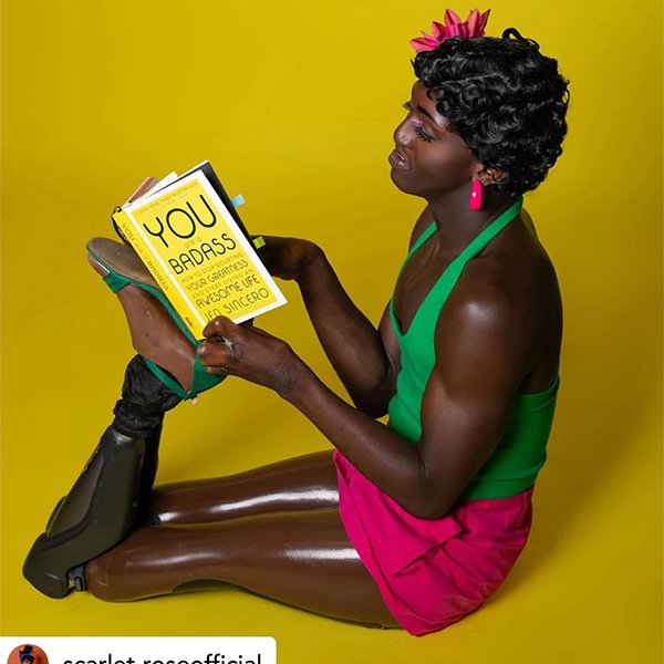 A person with a prosthetic leg sitting on the floor against a yellow background, holding a book titled 'You Are Badass'. They are wearing a green top, pink shorts, pink earrings, and a pink flower in their hair.