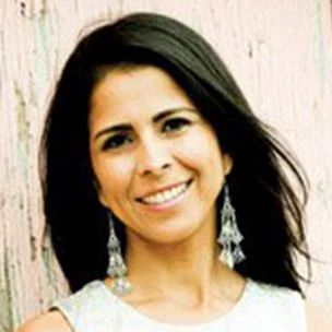 A woman with dark hair smiling, wearing earrings, standing in front of a light-colored wooden background.