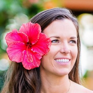 Woman with long brown hair and a large pink hibiscus flower behind her ear, smiling outdoors.