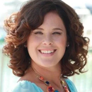 A woman with curly brown hair smiling outdoors on a sunny day.