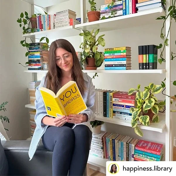 A woman with long brown hair and glasses sitting on a couch, reading a yellow book titled 'You Are Badass' in a room with white shelves filled with books and plants.