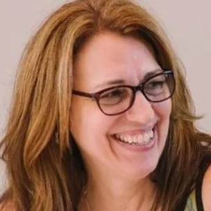 Close-up of a woman with long, reddish hair and glasses, smiling and looking to her right.