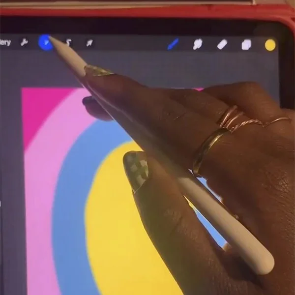 Close-up of a person's hand using a stylus on a tablet or touchscreen device with a colorful background. The person has rings on their fingers and is interacting with digital art or design software.