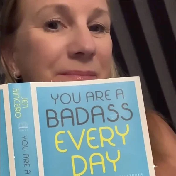 A woman holding a book with the cover reading 'You are a Badass Every Day' in front of her face, with part of her face visible, smiling softly.