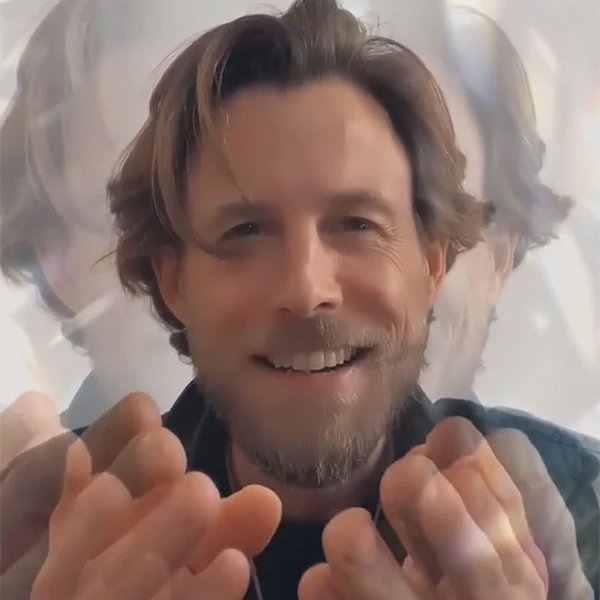 A smiling man with medium-length hair and a beard, making gestures with his hands, with a blurred background.