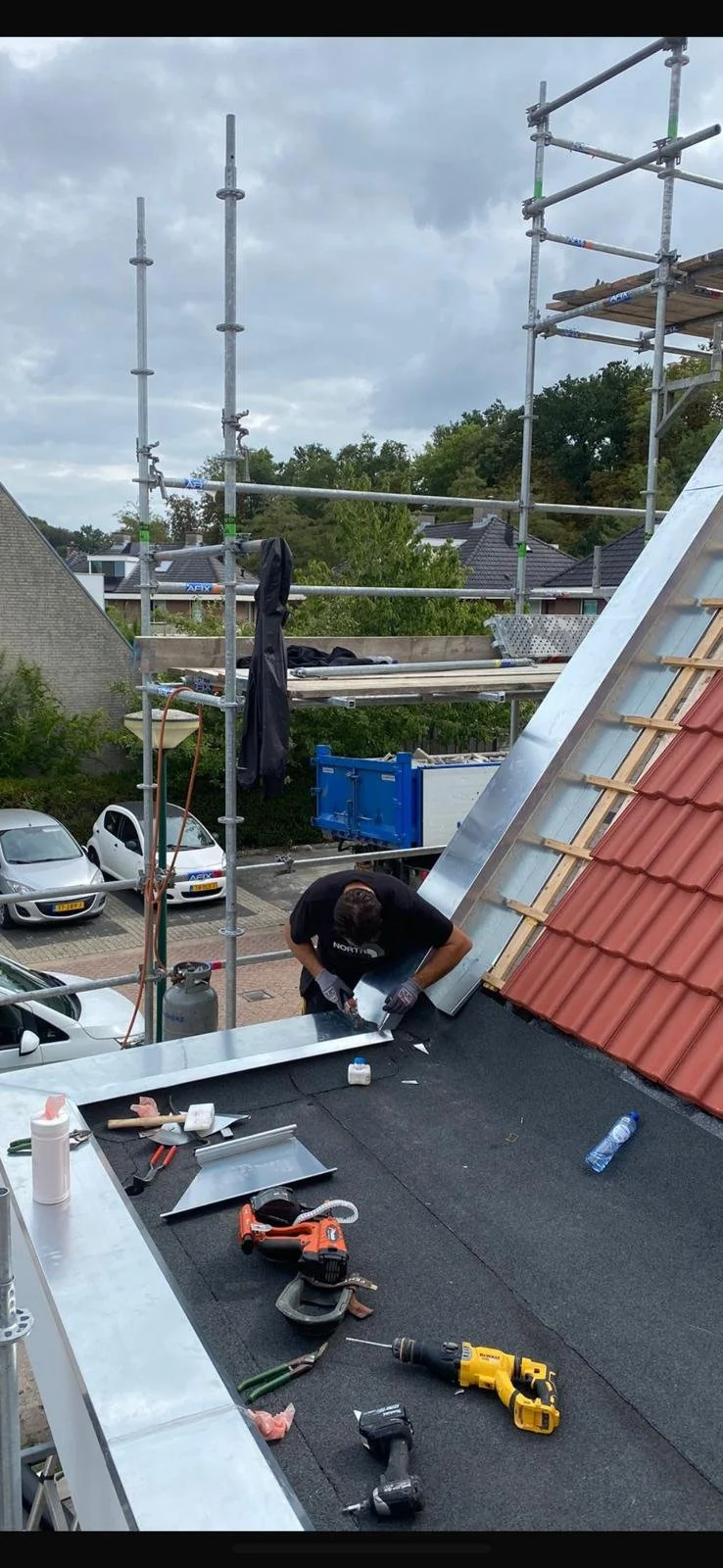 A construction worker installing metal flashing on a sloped roof with red tiles. Scaffolding is visible above, and tools such as a cordless drill, clamps, and screws are spread on the roof. Cars are parked below, and a cloudy sky is overhead.