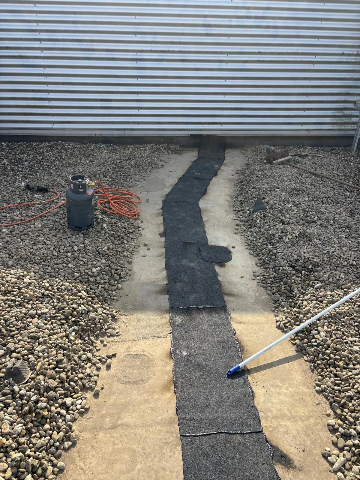 A pathway under construction with black asphalt patches on concrete, flanked by gravel and dirt, and a metal building in the background.