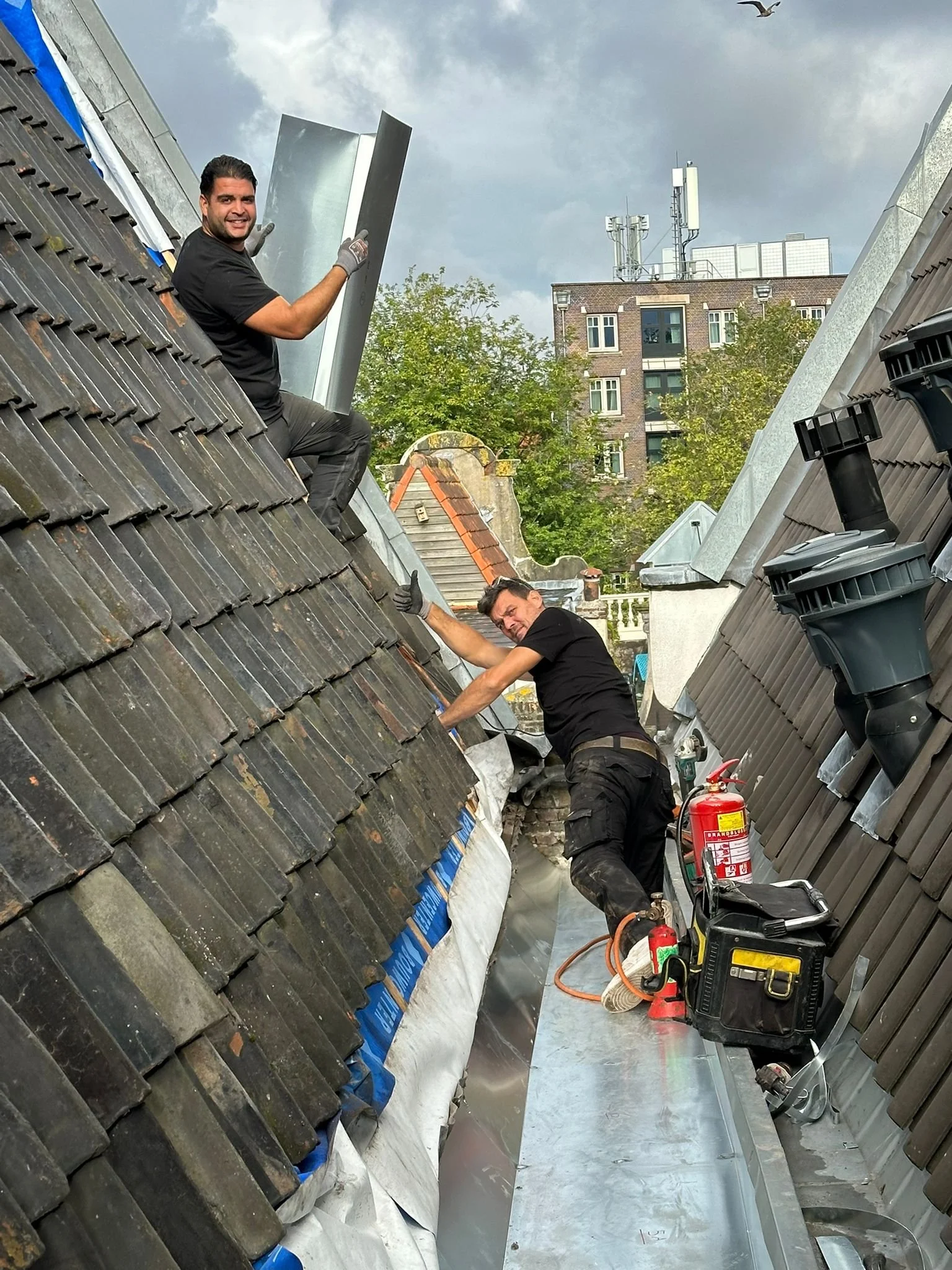 Two workers on a sloped rooftop installing or repairing roofing materials, with tools and equipment nearby, against an urban background with a cloudy sky.