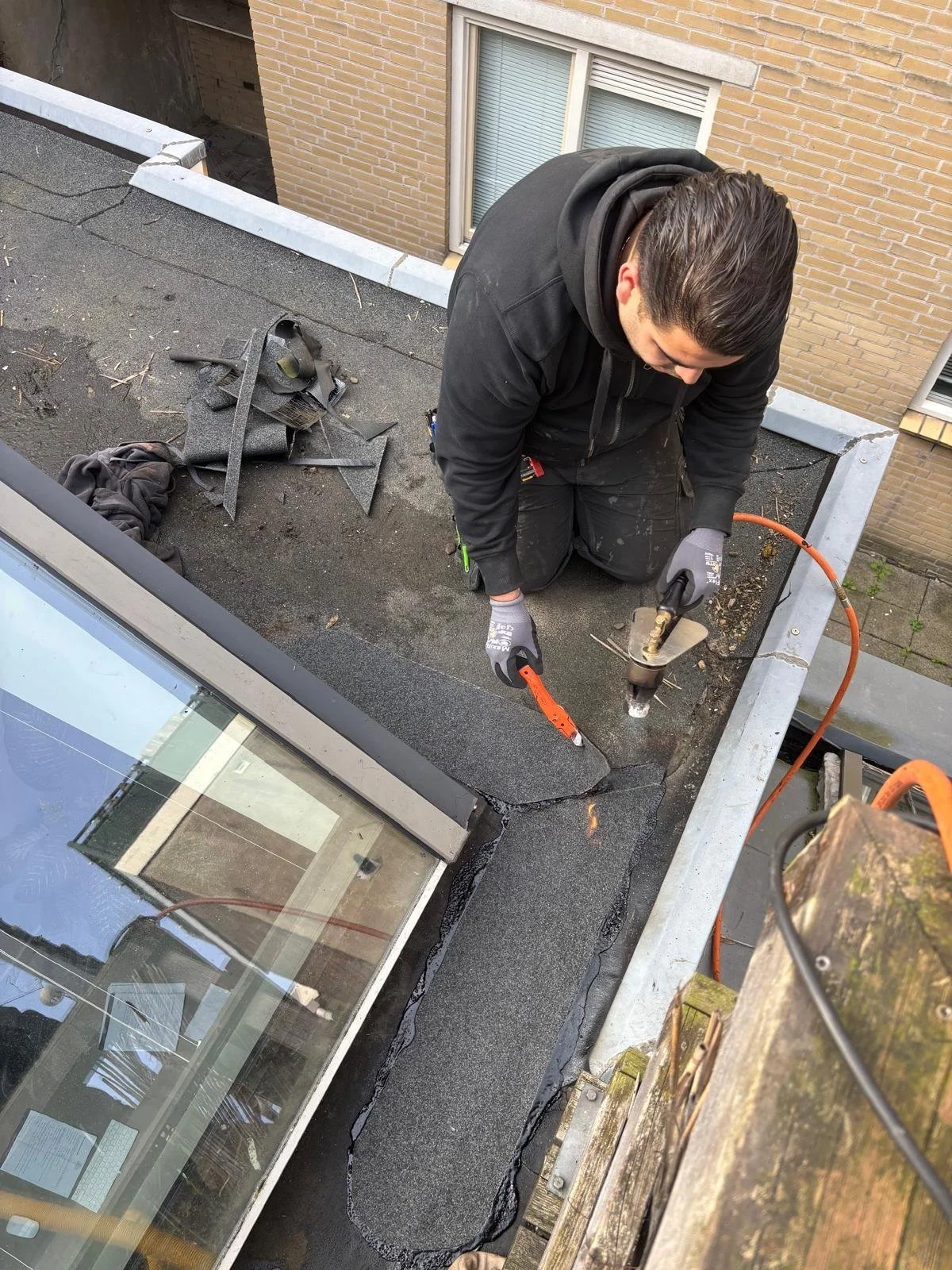A construction worker kneeling on a rooftop applying sealing material around a skylight window using a caulking gun. The worker is wearing gray gloves and a black hoodie. There are tools and scraps of roofing material nearby.