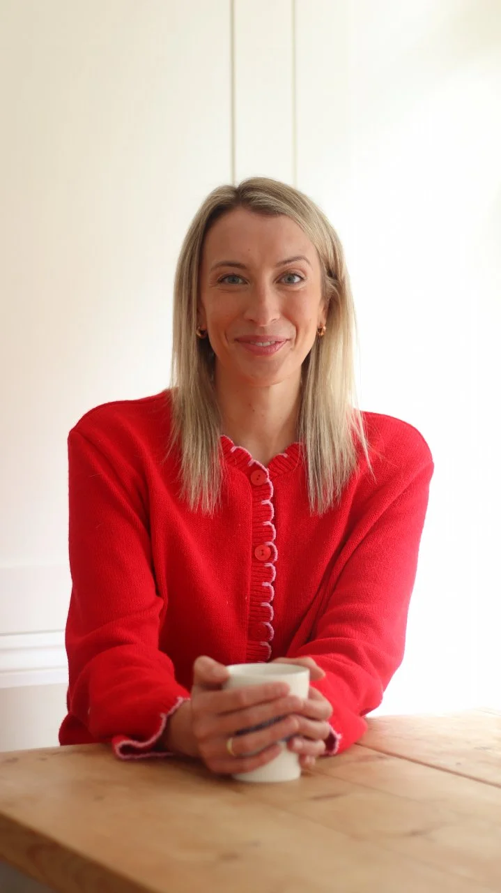 A woman with blonde hair, wearing a red sweater, sitting at a wooden table holding a white mug, smiling at the camera with a light-colored wall background.