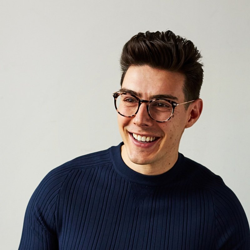 A young man with dark hair styled up, wearing round glasses and a navy blue ribbed shirt, smiling and looking off to the side against a plain white background.