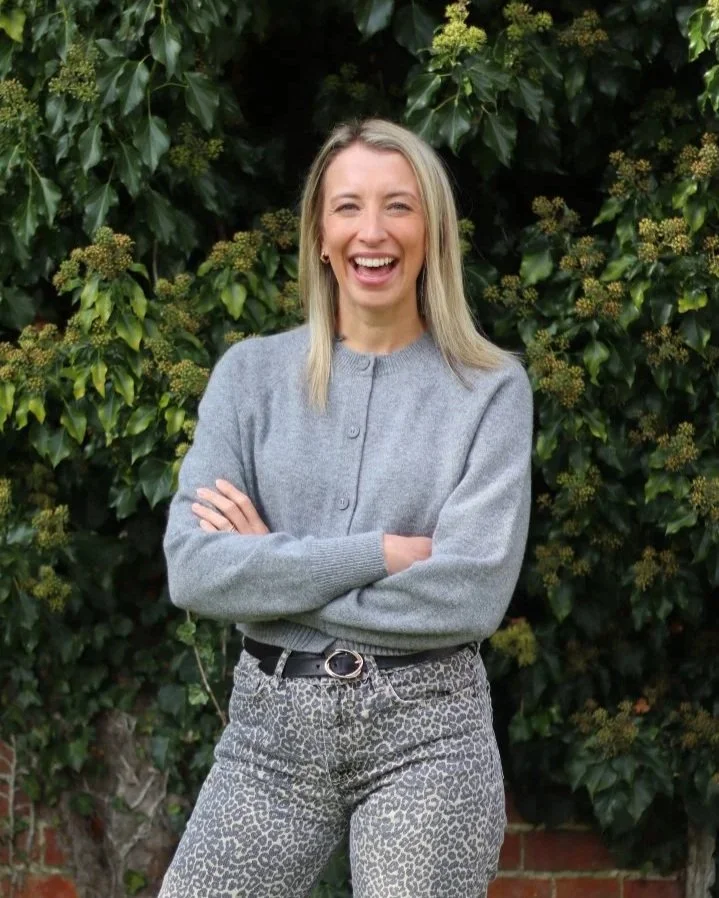A woman with blonde hair smiling and standing in front of green leafy bushes, wearing a gray cardigan and animal print pants.