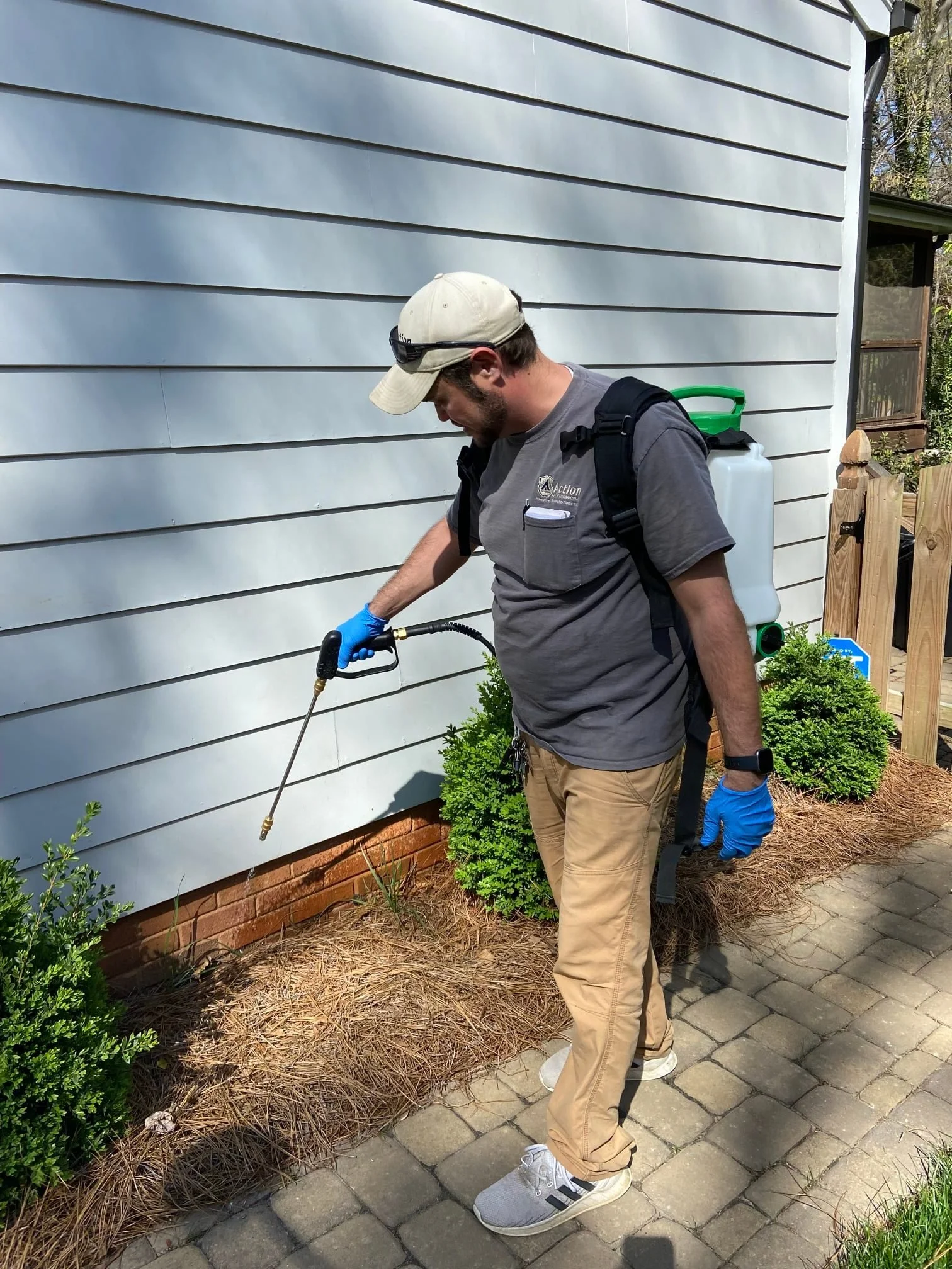 A man wearing a tan cap, gray shirt, tan pants, and blue gloves is using a sprayer on a shrub outside a house with gray siding, on a brick path.