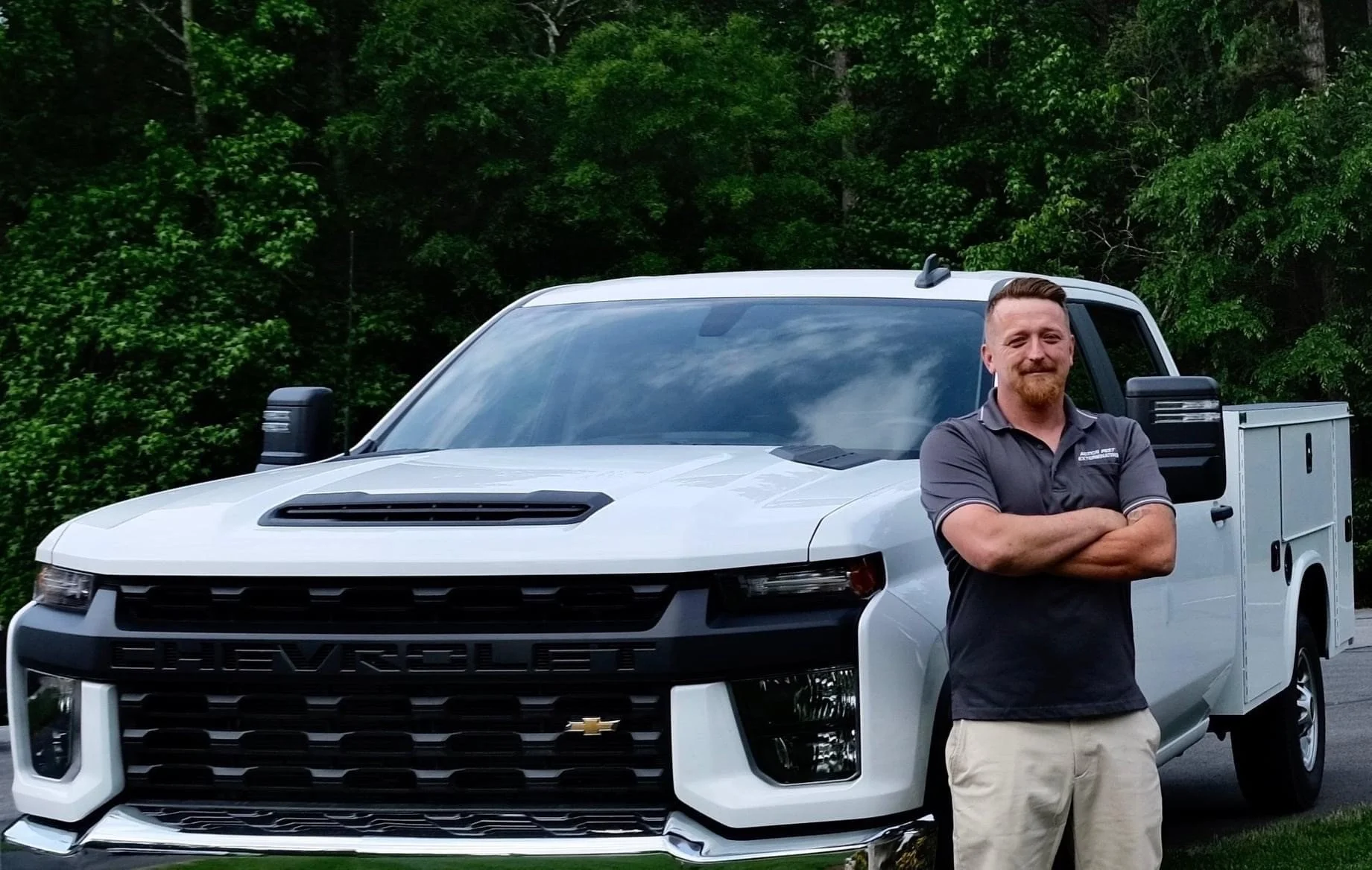 A man with crossed arms standing in front of a white Chevrolet Silverado truck with a Green background of trees.